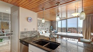 A kitchen with a granite countertop, a double sink, and a dishwasher. There is a dining area with a table and chairs, and a view of the outdoors through large windows.