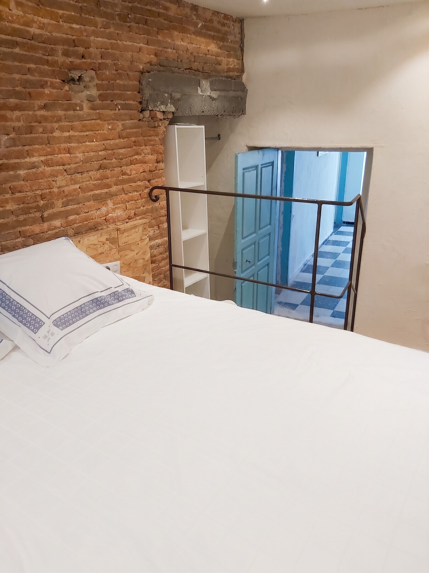 Une pièce avec un mur en briques, un cadre de lit en métal, un couvre-lit blanc et une porte bleue donnant sur un sol à carreaux.