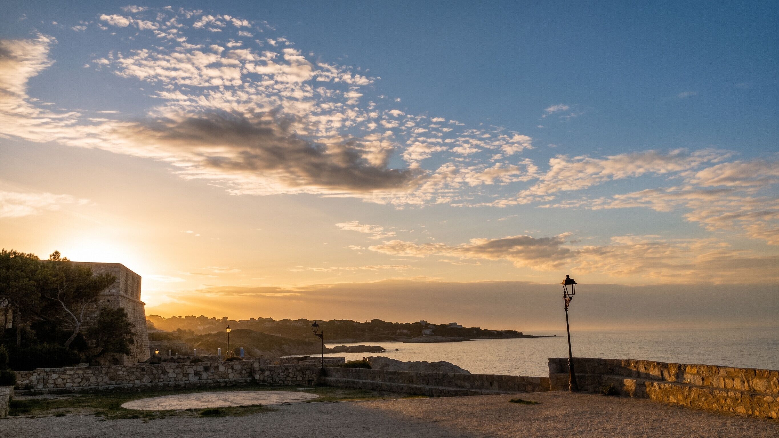 Sonnenuntergang am Meer mit Steinmauern und einer Straßenlaterne.