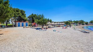 Uma praia com pedras, guarda-sóis e cadeiras de praia. Há árvores e um pequeno edifício com portas azuis.