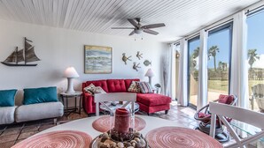 A spacious living room with a red sofa, a grey sofa, and a white coffee table. There is a ceiling fan, a large window with a view of palm trees, and a decorative sailboat on the wall. - Sandy Toes @ MP 116 | Pools | Beach Boardwalk (Port Aransas)