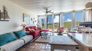 A spacious living area with a red sofa, a white sofa, and a dining table. There is a large window with a view of the outdoors. - Sandy Toes @ MP 116 | Pools | Beach Boardwalk (Port Aransas)