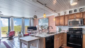 A modern kitchen with wooden cabinets, a black stove, and a large window overlooking a grassy area. - Sandy Toes @ MP 116 | Pools | Beach Boardwalk (Port Aransas)
