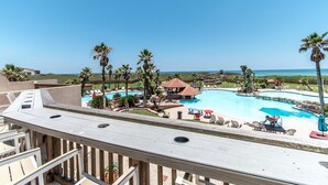 A resort pool area with palm trees, a wooden deck, and a view of the ocean. - Live Your Best Beach Life @ Mayan Princess 211 (Port Aransas)