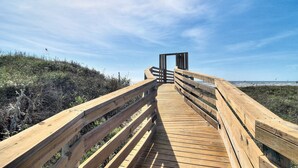 A wooden boardwalk leading to a structure with a glass door, surrounded by natural vegetation and a clear sky. - Live Your Best Beach Life @ Mayan Princess 211 (Port Aransas)