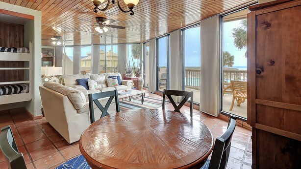 A spacious living area with a wooden ceiling, a round wooden table, and a large window offering a view of the outdoors. - Surf-Shells-Sandcastles | Ocean-Pool Views | MP214 (Port Aransas)