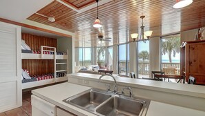 A kitchen with a double sink, wooden ceiling, and large windows offering a view of the outdoors. - Surf-Shells-Sandcastles | Ocean-Pool Views | MP214 (Port Aransas)