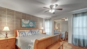 A bedroom with a wooden bed, two bedside tables with lamps, a ceiling fan, a bench, a large mirror, and a view into another room. - Surf-Shells-Sandcastles | Ocean-Pool Views | MP214 (Port Aransas)