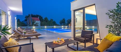 Uma área externa moderna com piscina, espreguiçadeiras e uma mesa de jantar com almofadas.
