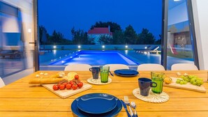 A dining table set with plates, cutlery, and a variety of food items, overlooking a swimming pool and a house in the background. - VILLA ALMIC with heated pool, 5 bedrooms, Media room,multi-use playground court (Naklice)