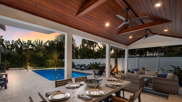 A covered outdoor dining area with a pool in the background, wooden ceiling, and outdoor furniture. - Take Me Away - Stylish Coastal Escape with Pool Near Beach and Trolley (Holmes Beach)