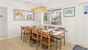 A dining area with a wooden table set for six, flanked by chairs, a hanging light fixture, and a window with blinds.
