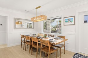 A dining area with a wooden table set for six, flanked by chairs, a hanging light fixture, and a window with blinds.