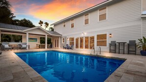 A swimming pool with a stone deck, a covered patio area, and a white building with large windows and a door. - Take Me Away - Stylish Coastal Escape with Pool Near Beach and Trolley (Holmes Beach)