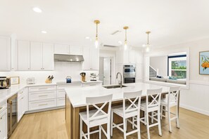 A modern kitchen with white cabinetry, a central island with bar stools, and pendant lighting. - Take Me Away - Stylish Coastal Escape with Pool Near Beach and Trolley (Holmes Beach)