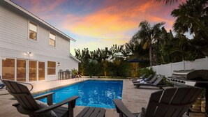 A swimming pool with lounge chairs and a grill area, surrounded by palm trees and a house. - Take Me Away - Stylish Coastal Escape with Pool Near Beach and Trolley (Holmes Beach)