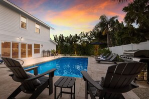 A swimming pool with lounge chairs and a grill area, surrounded by palm trees and a house.