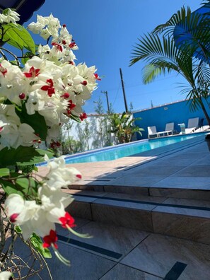 A pool area with clear blue sky, white flowers, and a blue building in the background. - Residencial Rosi (Caraguatatuba)