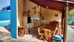 An outdoor patio with a barbecue grill, wooden table and benches, and a pool in the background. - Residencial Rosi (Caraguatatuba)