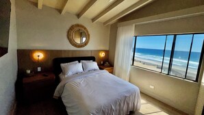 A bedroom with a bed, a ceiling fan, a round mirror, bedside lamps, and a large window with a beach view.