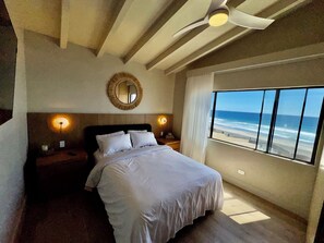 A bedroom with a bed, a ceiling fan, a round mirror, bedside lamps, and a large window with a beach view.