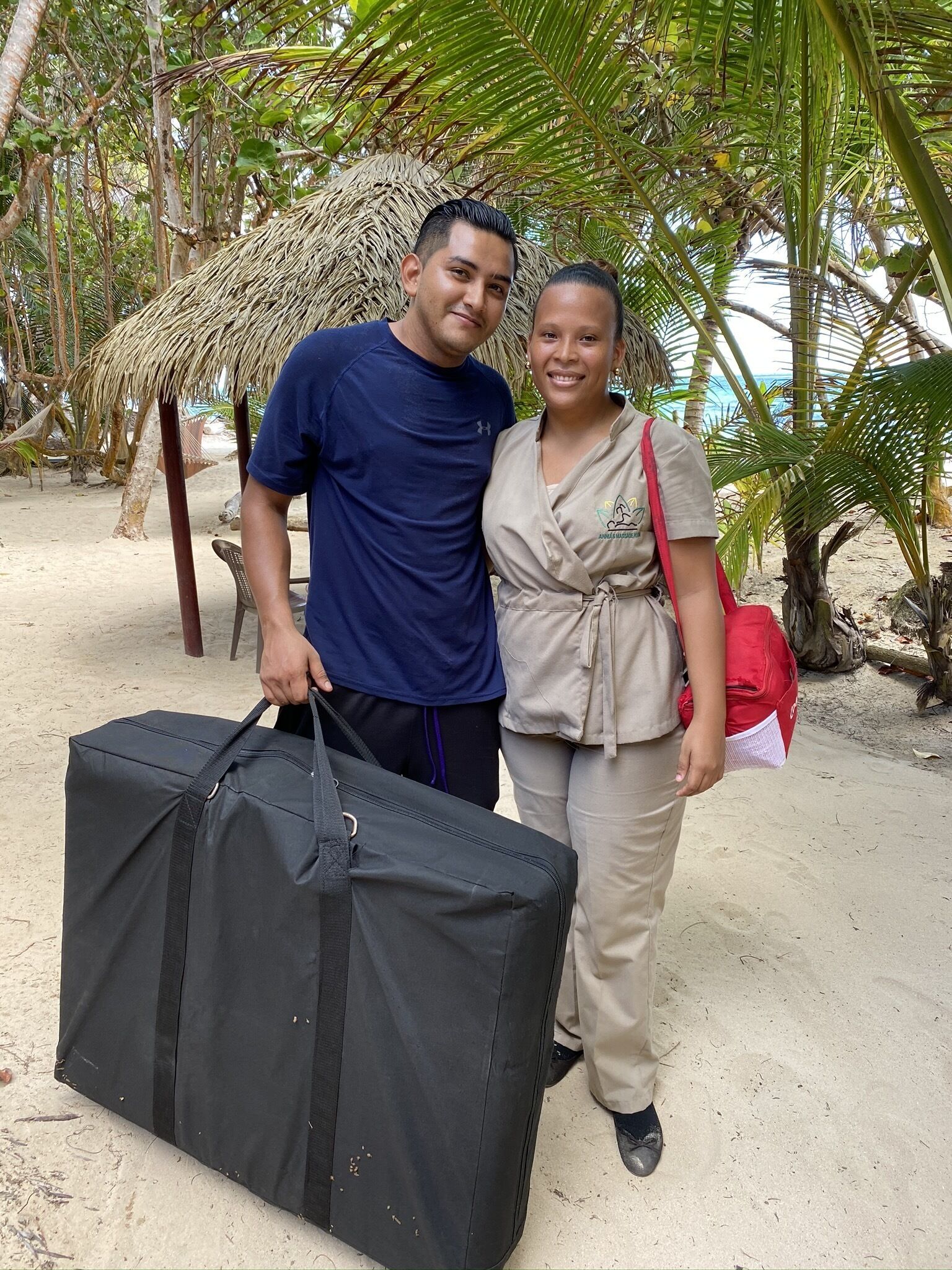 Two people standing on a sandy beach with palm trees and a thatched structure in the background.