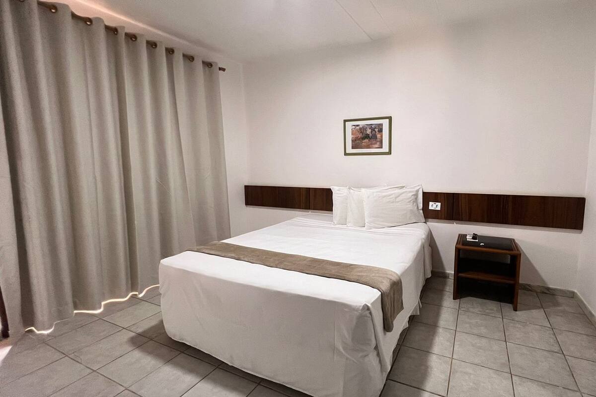 Quarto de hotel com cama, mesa de cabeceira, um quadro na parede e janela com cortinas.