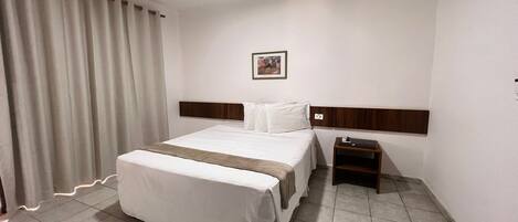 A hotel room with a bed, a nightstand, a picture on the wall, and a window with curtains.
