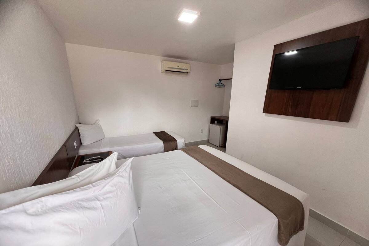 Quarto de hotel com duas camas de solteiro, televisão fixada na parede, ar condicionado e uma pequena escrivaninha com cadeira.