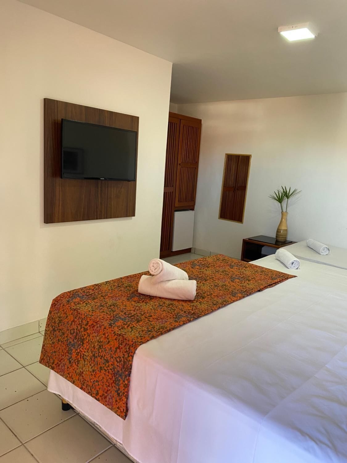 A hotel room with a bed, a flat-screen TV mounted on the wall, a wooden wardrobe, a small table with a vase, and a potted plant.