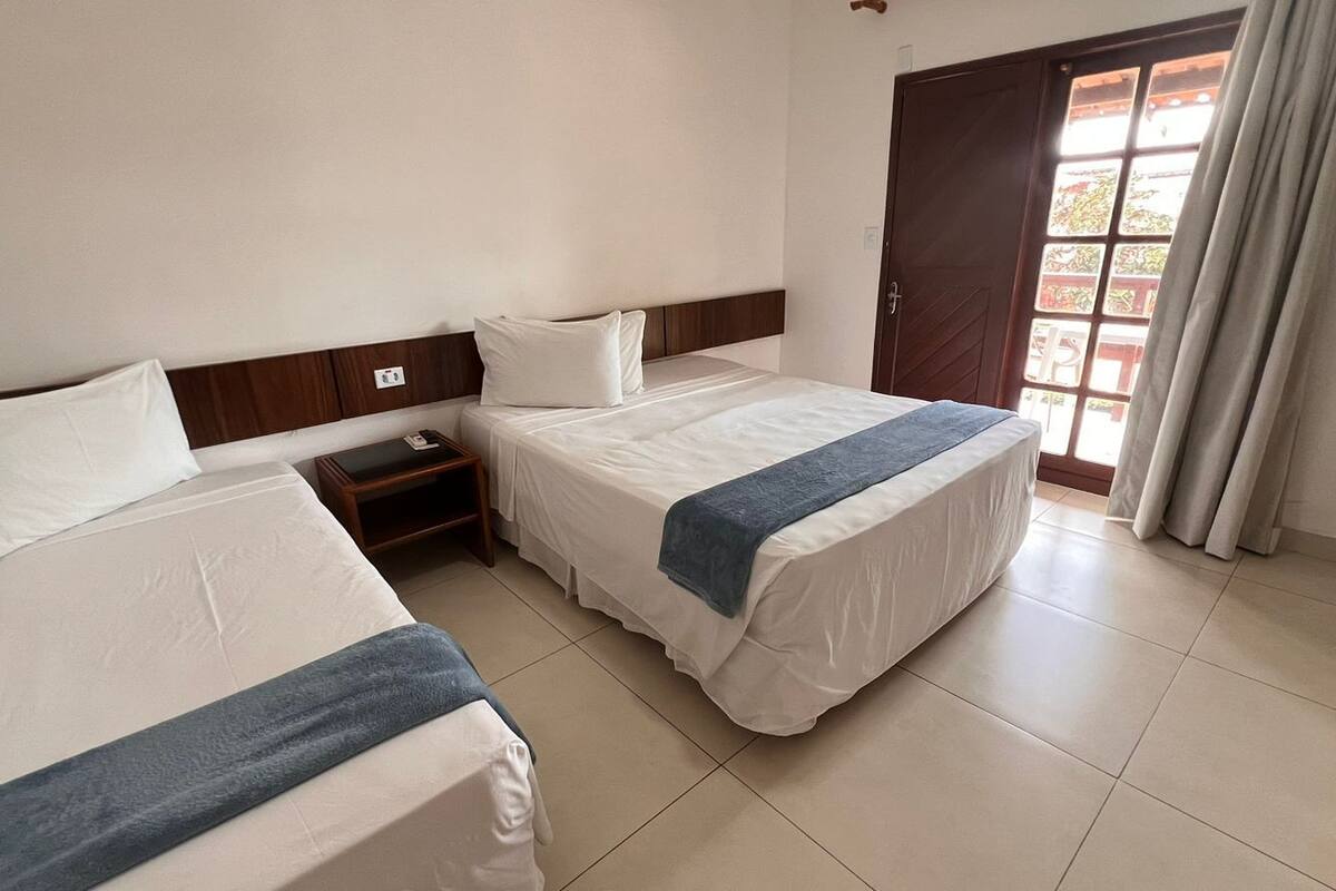 Quarto de hotel com duas camas, cabeceira de madeira, mesa de cabeceira pequena e janela com cortinas.