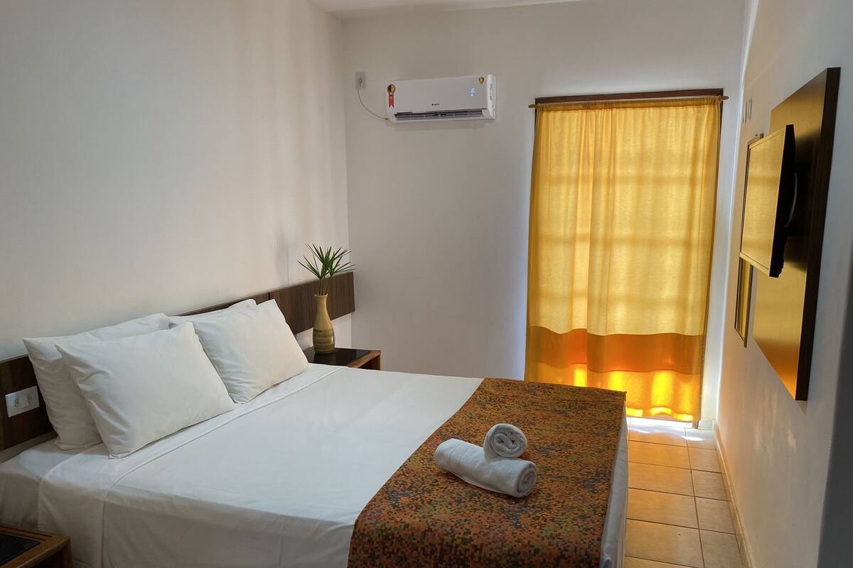 Quarto de hotel com cama, mesa de cabeceira, ar condicionado, janela com cortinas e televisão na parede.