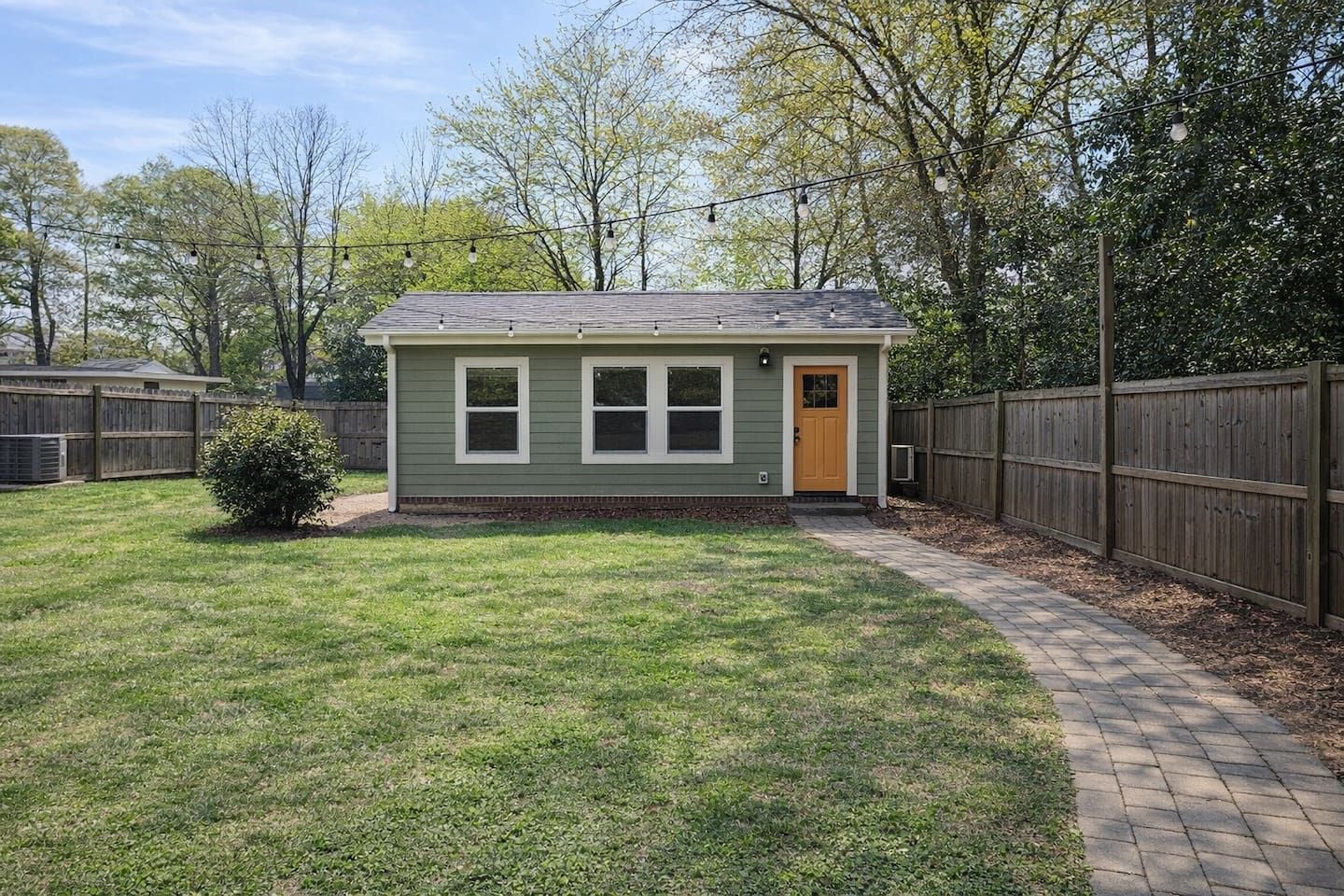 A small, single-story house with a front door and windows, surrounded by a wooden fence and a paved walkway.