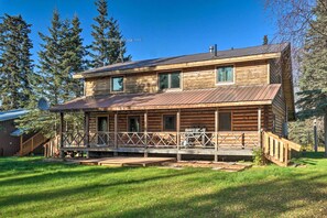 Eine zweistöckige Blockhütte mit einer Veranda und einer Terrasse, umgeben von einem Rasen und Bäumen.