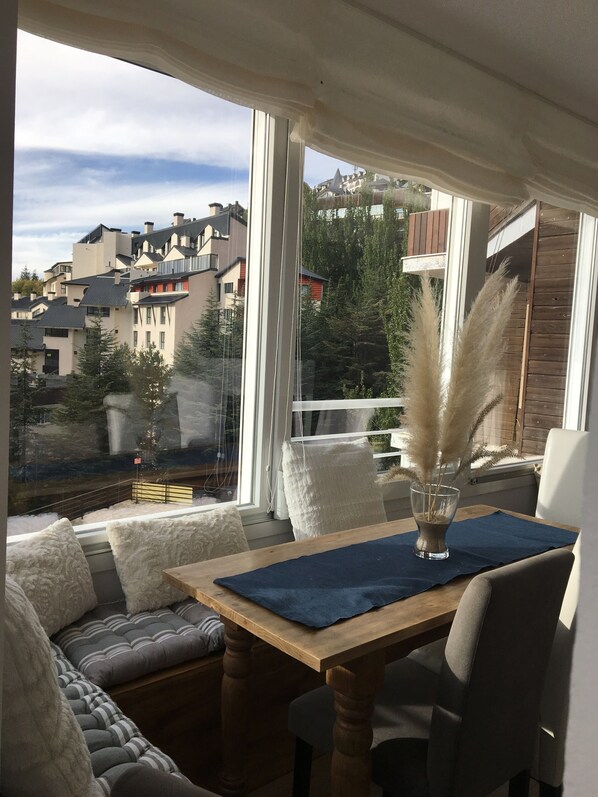 Un rincón acogedor con una mesa de madera y sillas, un jarrón con plumas de pampa y vistas a un barrio residencial a través de amplias ventanas.