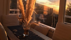 A dining area with a table set for two, a vase with pampas grass, and a view of the cityscape through large windows.