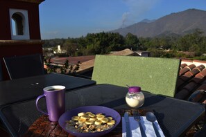 A table with a purple mug, a bowl of cereal, and a small jar of yogurt, set against a backdrop of a mountain and trees. - Casa San Miguel Arcangel (Antigua Guatemala)
