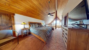 Une chambre avec un plafond en bois, un lit avec une literie à motifs, une table de chevet avec une lampe, une commode et un téléviseur.