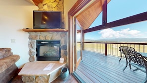 A stone fireplace with a television above it, a wooden table with a menu, and a deck with a view of the landscape.