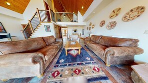 A spacious living room with two brown leather sofas, a wooden coffee table, and a patterned rug. A staircase with a wooden railing leads to an upper level.