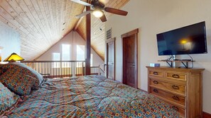 A bedroom with a sloped ceiling, a bed with patterned bedding, a wooden dresser, a television, and a balcony with a railing.