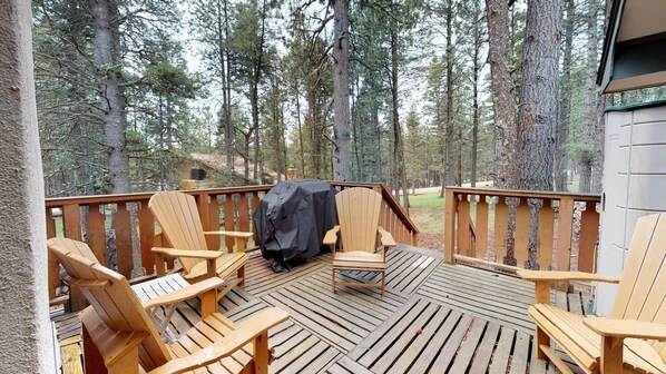 Een houten terras met Adirondack-stoelen en een barbecue, omgeven door hoge bomen.