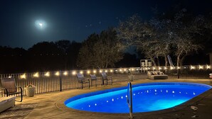 A swimming pool at night with a fence, lounge chairs, and string lights.