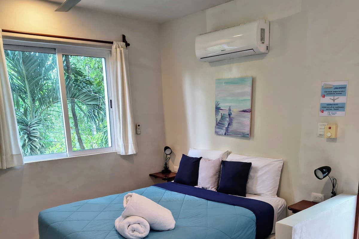 Un dormitorio con cama, ventilador de techo, aire acondicionado, ventana con cortinas y un cuadro en la pared.