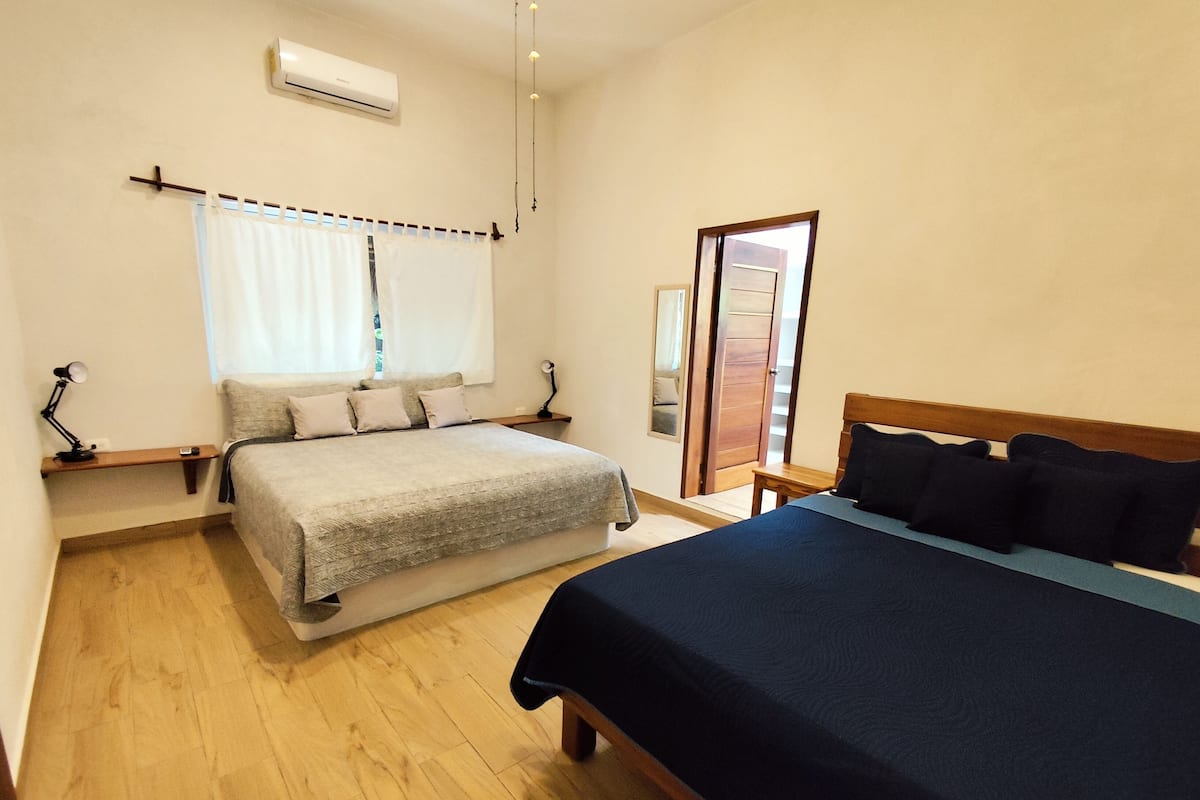 Un dormitorio con cama, mesita de noche, lámpara, ventana con cortinas y aire acondicionado.