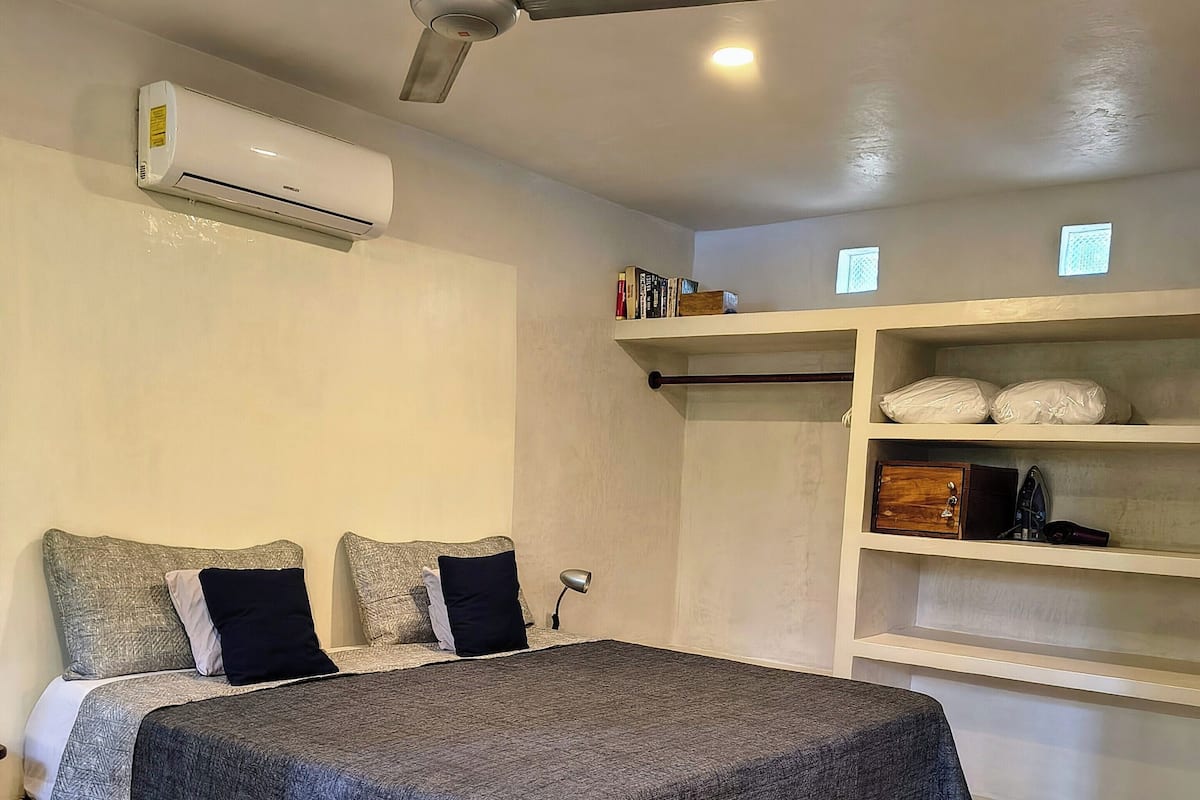 Un dormitorio con cama, ventilador de techo, equipo de aire acondicionado, lámpara de noche y estantes empotrados con objetos encima.