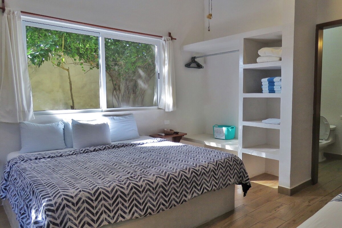 Un dormitorio con cama, ventana con vistas a árboles, baño con lavamanos y espejo, y estante con toallas.