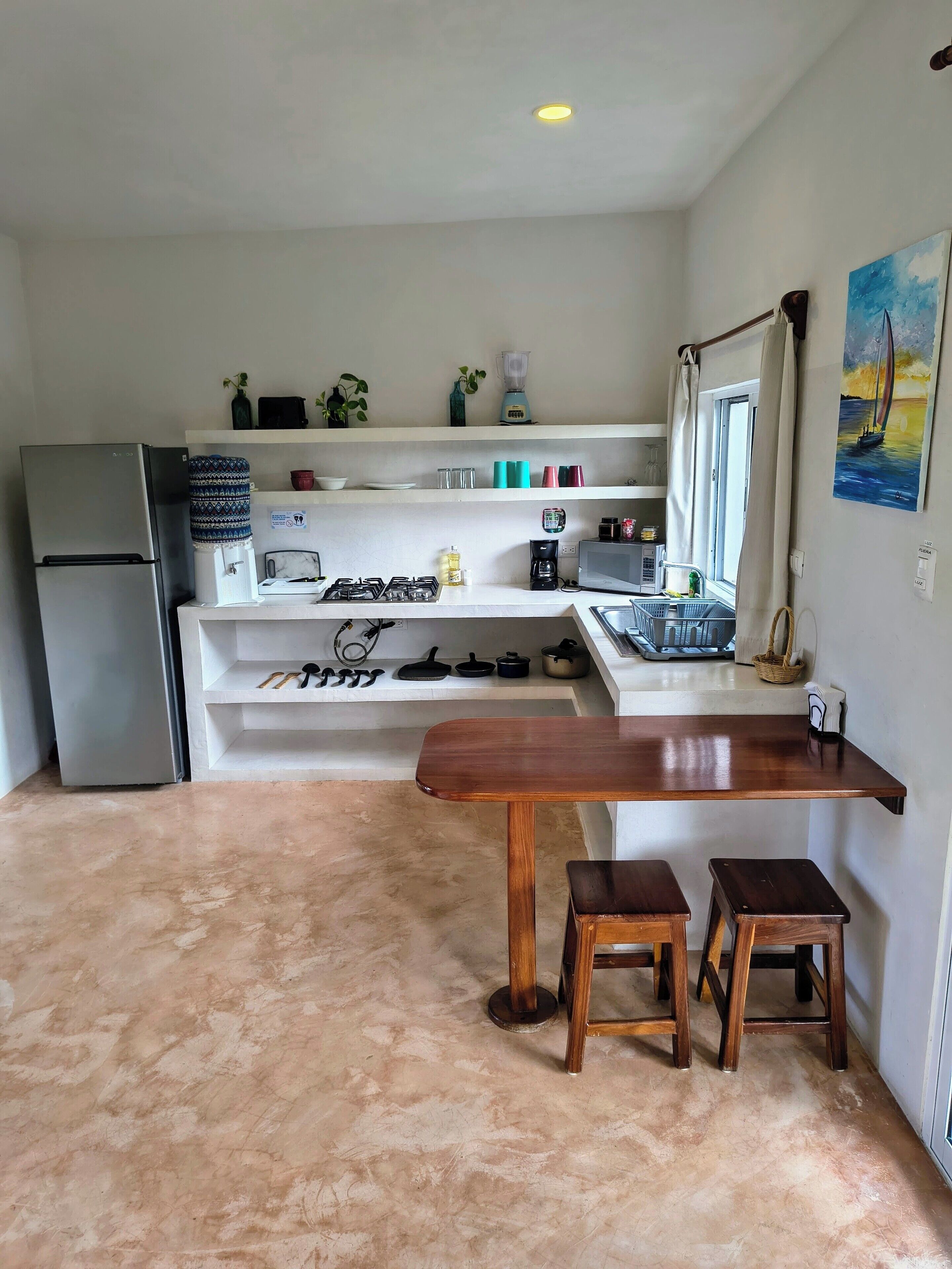 Una cocina con refrigerador, estufa y mesa de madera con taburetes. Hay repisas sobre la encimera con diversos objetos.