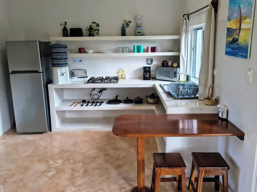 Una cocina con refrigerador, estufa y mesa de madera con taburetes. Hay repisas sobre la encimera con diversos objetos.