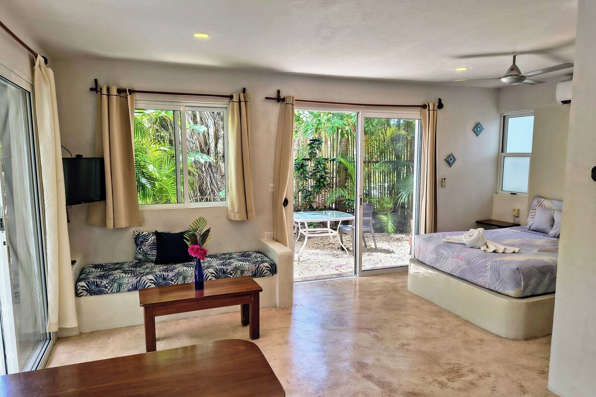 Una habitación con una cama, un banco, una mesa, un televisor y vistas a un jardín a través de puertas correderas.
